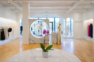 Bright and airy interior of the Jendue luxury denim boutique at Town Center Boca Raton Mall, featuring a pink orchid in a white pot on a round marble table in the foreground. In the background, a circular illuminated marble display panel frames gold manne