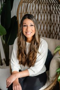 Stacy Padula smiling warmly while seated in a wicker egg chair, wearing a white blouse with long wavy brown hair, surrounded by green houseplants.