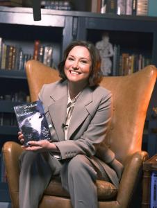 Actress Chyler Leigh sits in a high-backed leather library chair, smiling and holding a copy of In the Shadow of a Rainbow.