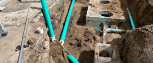 A commercial plumbing construction site featuring deep excavation trenches with heavy-duty green sewer piping, concrete junction boxes, and cleanout access points installed by KPS Plumbing.