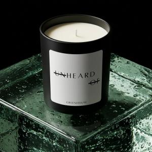 A signature matte black UNHEARD OF candle labeled "Greenhouse" sitting on top of a vibrant, bubbly emerald green glass block against a dark background.