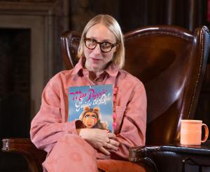Author Emma Straub poses in a high-backed leather library chair, holding a copy of Miss Piggy's Guide to Life.