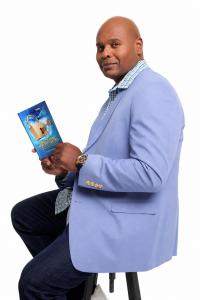 Kevin D. Williams seated holding his bestselling book Unlocking Opportunities in light blue blazer, Dallas Communication Strategist and Certified World Class Speaking Coach.