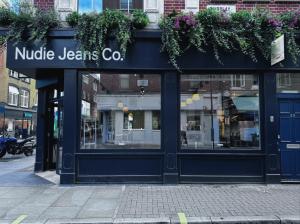 Exterior of Nudie Jeans Co. store in Soho, London. Navy blue shopfront with large glass windows, white Nudie Jeans Co. signage, and trailing green plants and purple flowers above the entrance. Number 29 visible on the right hand side.