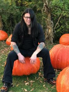 Author David Grimsby sitting on a large pumpkin in an outdoor pumpkin patch, wearing black clothing and glasses, surrounded by oversized orange pumpkins and autumn foliage.