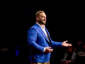 Phil Neil, founder of Founders Compass, on the TEDxHarvard Square stage in a blue plaid blazer, gesturing with both hands open while delivering his talk "When Founders Break, Businesses Follow."