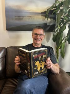 Image of author Jeff Wright, a smiling middle-aged man with short greying hair and wearing glasses, holding a hardcover copy of his new book, The Full Disclosure Iliad..