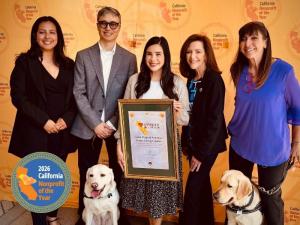 4 women and one man, along with 2 yellow labradors standing in front of orange backdrop with CA Nonprofit logos printed on it, holding up a frame with certificate for being recognized as a 2026 California Nonprofit of the Year.