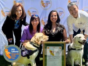 3 women and 1 man kneeling with framed 2026 California Nonprofit of the Year award, with 2 yellow Labrador retrievers in the foreground.