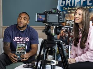 Two people sit side-by-side during a book promotion or interview. On the left, a man wearing a navy blue "JESUS WON" t-shirt holds a copy of the book Gripped by Stacy A. Padula. On the right, a woman with long brown hair in a pink hoodie smiles at the cam