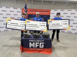 Three young men wearing blue shirts stand behind a table at a Project MFG awards event, each holding oversized prize checks for a maritime welding competition. The center winner holds a $10,000 first-place check, while the others hold $5,000 second-place