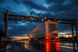 A commercial semi-truck passing under an automated electronic compliance gantry and toll scanners on a wet highway at twilight, representing commercial freight logistics and the digital monitoring of the American trucking industry.