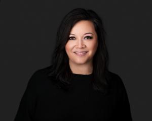 A woman with shoulder-length dark hair smiles at the camera during a professional headshot session. She is wearing a black sweater against a dark gray studio background.