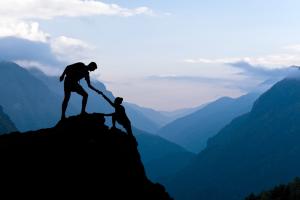 photo schema, black mountain with blue sky with man on top of mountain reaching his hand down and helping a man climb up the mountain