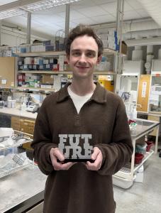 Image of Foundation Fellowship winner, a young man, with short brown hair, standing in a laboratory holding his prize