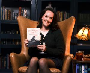 Jena Malone sits in a high-backed leather library chair, holding a copy of The Flowering Wand by Sophie Strand
