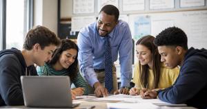 Male high school teacher leaning over a table while assisting a diverse group of students engaged in classroom learning and discussion.