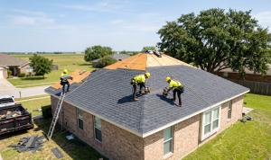 Roofing crew working on a residential home, installing shingles on a sunny day with clear skies and green surroundings.