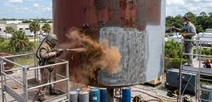 Two workers in protective gear sandblast and paint a large, rusted metal tank. One sprays sand, while the other paints. Industrial setting, clear day.