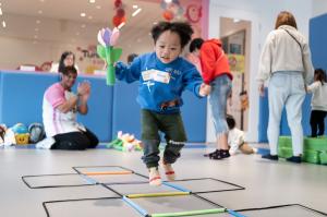 一名幼兒在 TuniTuni 故事體能課堂中，手持花朵造型教具跳過彩色地面梯。