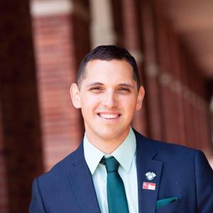 A White male in a navy suit, with a navy-blue tie, light blue shirt, with a navy haircut, smiling.