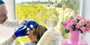 Dual board-certified Dr. Zarrini administers a precision facial injection on a patient inside a bright Beverly Hills medical spa overlooking palm trees.
