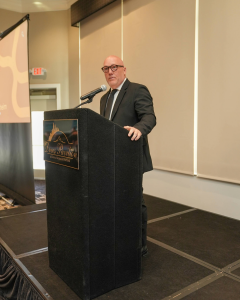 OneQuest Health Board President Jeffrey Sackenheim stands behind a podium addressing an audience at the OneQuest Health annual breakfast.