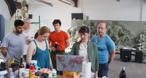 Traci L. Slatton stands at a laptop in studio with Master Sculptor Sabin Howard, sculptors and models gathered around her. Behind them are large clay figures from A Soldier’s Journey, with studio lights, tools and worktables visible.