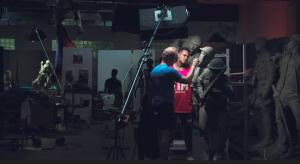 Sabin Howard sculpts a clay World War I soldier figure in a dim studio while a live model poses nearby. Lighting equipment, tools, and unfinished monument figures surround them during work on A Soldier’s Journey for the National WWI Memorial.