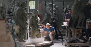 Sabin Howard sits on the studio floor working on a clay sculpture while assistant sculptor Charles Mostow stands nearby. Large unfinished figures from A Soldier’s Journey surround them during creation of the National WWI Memorial bronze relief.