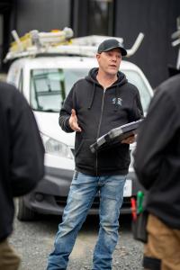 Steeve Plouffe, président d'Air Solution Confort, photographié sur un chantier en pleine explication, geste de la main à l'appui, tenant une mallette à outils robuste sous le bras. Il porte la casquette et le coton ouaté noir aux couleurs de l'entreprise.