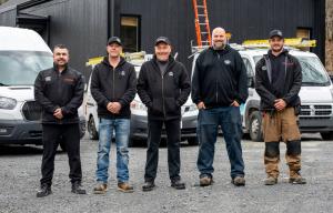 Photo de groupe réunissant cinq membres de l'équipe d'Air Solution Confort, tous vêtus de cotons ouatés ou manteaux noirs à l'effigie de l'entreprise, certains affichant les logos des partenaires SHARP et GREE. L'équipe pose fièrement devant trois camionn