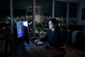 A person working late at a desk illuminated by blue and violet monitor light with a city skyline visible through the window behind them.