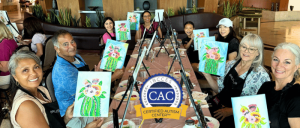 Group of people at a painting class, each holding a canvas with a colorful cactus and flower artwork, seated around tables with easels.