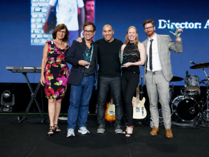 A group of celebrants stand on a stage celebrating an award for the Anthem Film Festival. Pictured are two women and three men.
