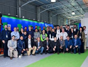 A group of professionals from the Brazilian delegation of Brazil Machinery Solutions poses for a group photo inside an international trade fair venue, with the booth in the background featuring blue and green tones, an industrial pavilion structure, and s