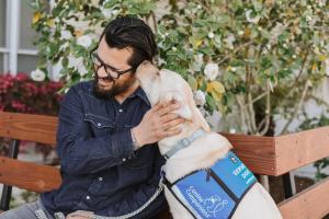 A smiling man is seated on a bench beside his service dog that is licking the side of his face.