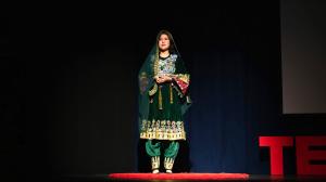 A woman wearing a traditional green embroidered outfit stands on a stage speaking during a TEDx presentation, with a dark backdrop and red TEDx letters visible beside them.