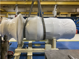 Large industrial equipment component fully enclosed in industrial shrink wrap inside a manufacturing facility prior to shipment.