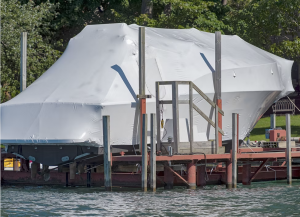 Large boat completely enclosed in white industrial shrink wrap while stored on a waterfront lift.