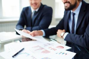 Deux conseillers en costume-cravate consultent ensemble des documents financiers couverts de graphiques et de tableaux statistiques, dans un bureau lumineux. La scène évoque l'analyse de données et la collaboration professionnelle dans le secteur des serv