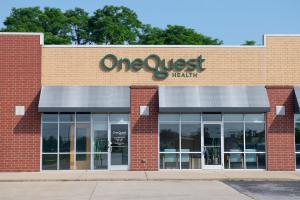 The exterior of the OneQuest Health outpatient facility in Grant County. The building has a modern facade combining red and tan brickwork. A dimensional green sign reading 