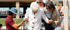 A young man and a middle-aged woman assist an elderly woman in a white coat as they hold hands outdoors in a sunny setting.