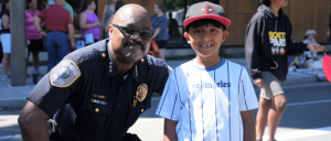 Police officer kneeling next to a smiling boy wearing a Deadpool hat and a Los Angeles baseball jersey at an outdoor event.