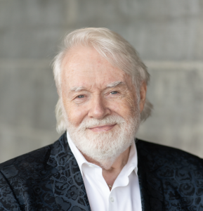 Headshot of Alan W. Green smiling at the camera. He has white hair and a white beard and is wearing a dark blue patterned blazer with a white shirt. The background is softly blurred gray.