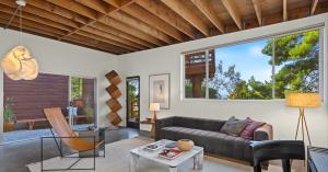 Modern living room in The Benson House, designed by Frank Gehry, with exposed wooden ceiling, gray sofa, marble coffee table, leather lounge chair, and large window showing trees. Warm lighting and minimalist decor create a cozy, contemporary atmosphere.
