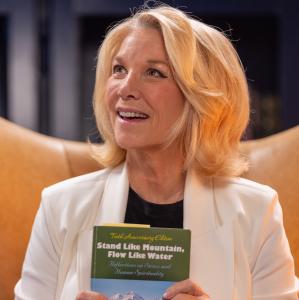 Joan Lunden sits in a leather high-backed chair, smiling and holding her copy of "Stand Like Mountain, Flow Like Water" by Brian Luke Seaward.