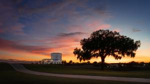 The Club at Iron Lake clubhouse at sunset in Ocala, Florida, with a large oak tree in the foreground, a golf course and cart path, and a colorful evening sky at IronCrest, a master planned gated lifestyle community.