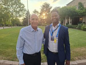 US Ambassador to Türkiye Tom Barrack, wearing a light blue shirt, stands beside I News Australia founder Nelly Yoa, who wears a navy suit and a NATO Summit press credential, on a lawn outside a building in Ankara at sunset.