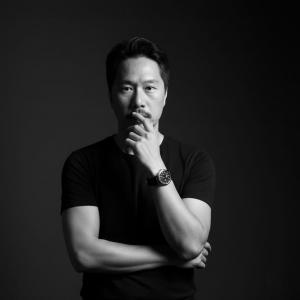 Portrait of David Johnson Jiang, a middle-aged man in a black T-shirt, standing with arms crossed and a thoughtful expression in a studio-style black background.